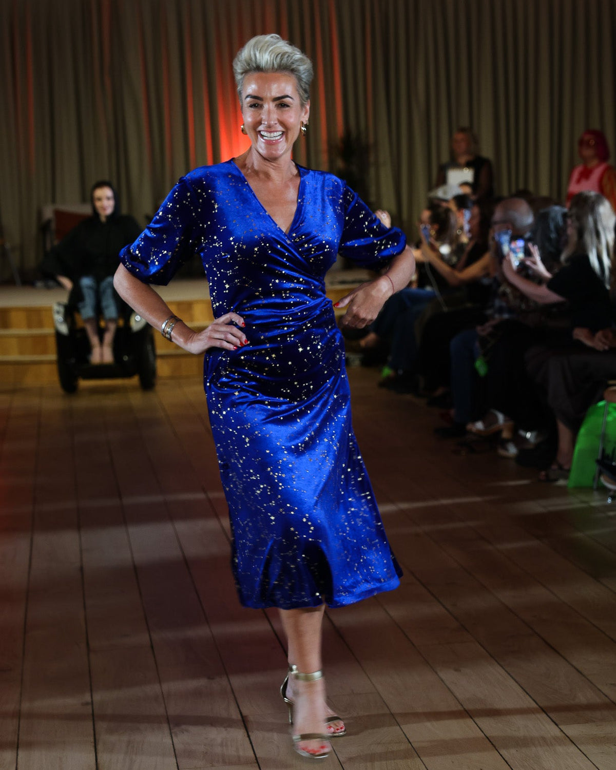 A woman models a sparkling royal blue embroidered with stars midi wrap dress with short puff sleeves and a V-neckline, walking on a wooden runway with an audience seated in the background.