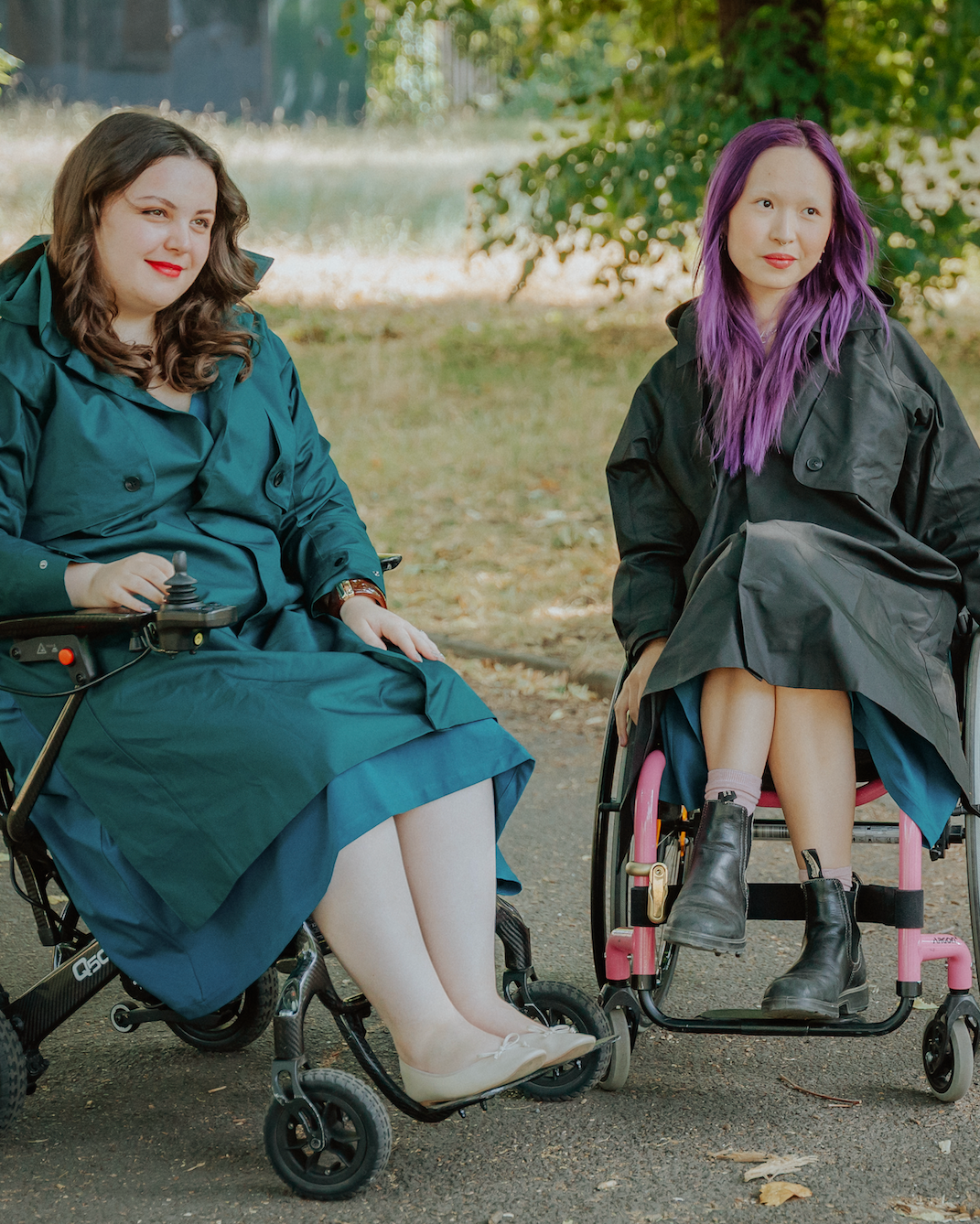 Two women sitting in wheelchairs outdoors on a paved path near a tree. The woman on the left wears a teal coat and skirt, and the woman on the right wears a black coat and skirt with bright purple hair.